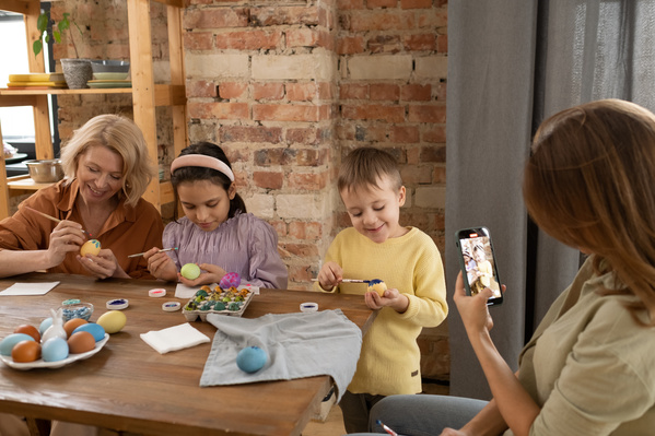 A woman takes a picture of her family consisting of two children and their grandmother painting eggs for Easter at a wooden table