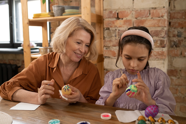 A woman with blonde hair holding a decorated Easter egg and a brush looks at her granddaughter decorating one of the eggs