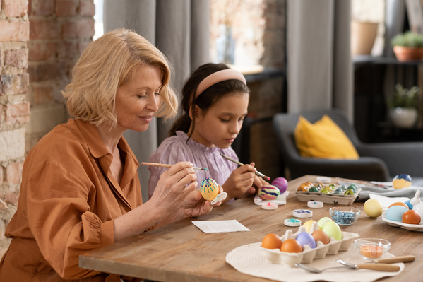 A blonde woman in a dress and her granddaughter with a pink headband on her head paint Easter eggs sitting at a table