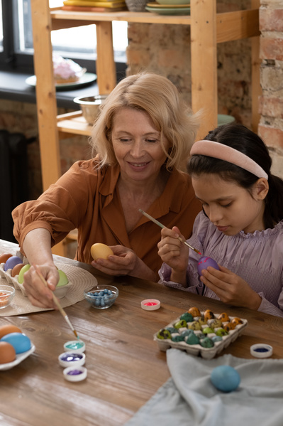 A girl in a pink blouse and her grandmother with blonde hair painting Easter eggs sitting at a wooden table with coloring materials
