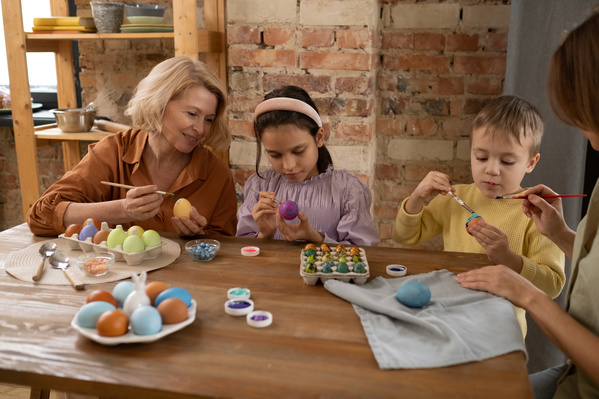A grandma with blonde hair holding an egg and a brush in her hands watches her grandchildren painting Easter eggs sitting at a wooden table on which painted eggs