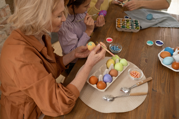 Granny with blonde hair drawing a blue flower on an Easter egg sitting at a wooden table with egg cartons