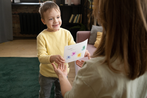 A boy with brown hair dressed in a yellow sweater presents an Easter card decorated with flowers and congratulations to his mother
