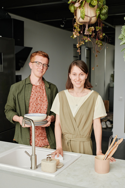 A man with red hair wearing a dark green shirt holding a stack of plates and a woman with short dark hair are in the kitchen