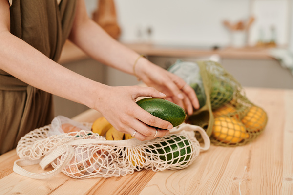 A woman with lays out an avocado from a string bag filled with vegetables and fruits on a wooden table