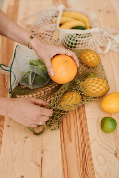 A woman puts citrus fruits from a string bag filled with vegetables and fruits on a wooden table