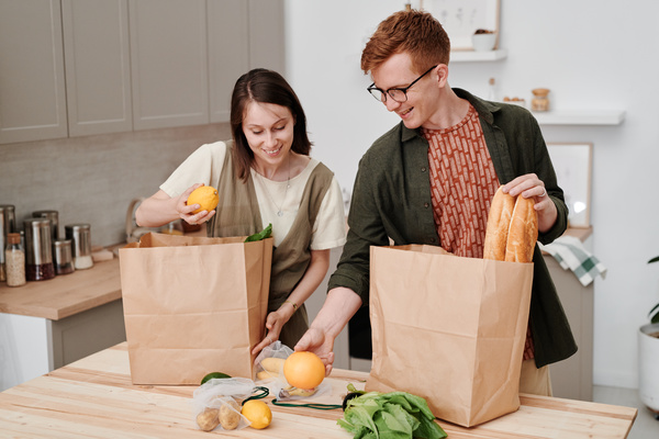 A couple of smiling young people sorting out grocery purchases from paper bags standing on a table with fruits and vegetables
