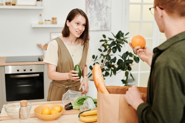 A woman with an avocado in her hand standing at a table with fruits and vegetables talks to a man laying out purchases from a paper bag