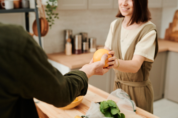 A man in a green shirt passes a grapefruit to a woman with short dark hair in the kitchen