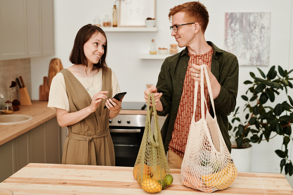 A man with red hair dressed in a dark green shirt holds white and green cotton string bags with groceries standing in the kitchen with a woman with short dark hair with a smartphone in her hands