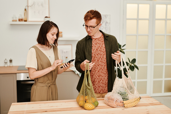 A guy with red hair dressed in a dark green shirt holds grocery in a string bag standing in the kitchen with a woman with a square with a smartphone in her hands