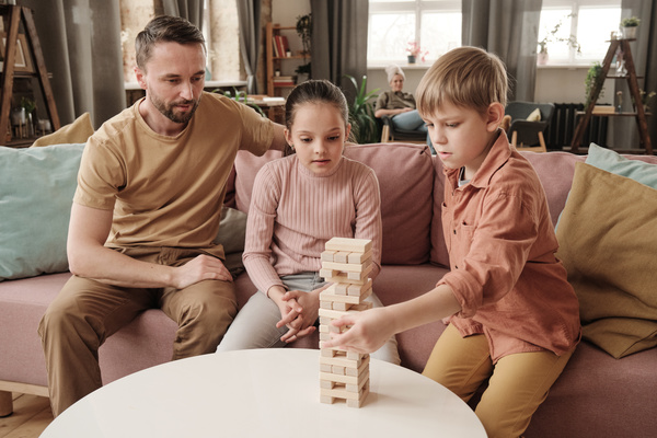 A boy with blond hair takes out a wooden block from a jenga playing with his sister and his father while sitting on the couch