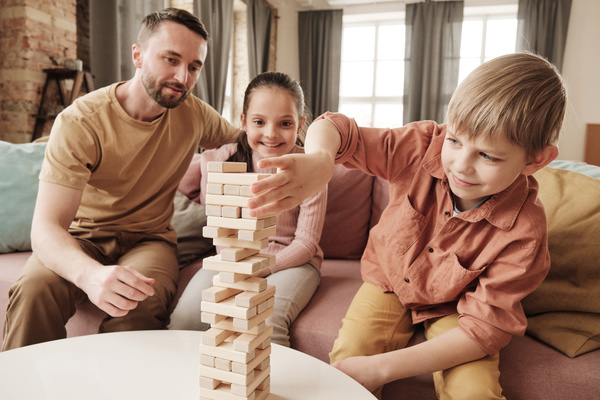 A boy with blond hair plays jenga with his sister and father sitting on a pink sofa
