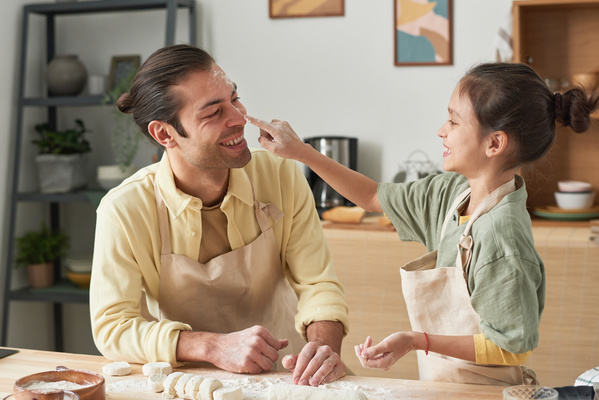A girl with covered in flour finger touches the nose of her father in an apron leaning on the kitchen table with dough and flour on it