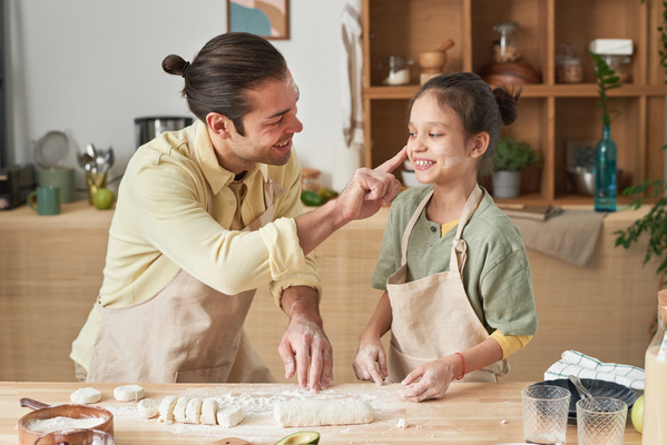 A father in an apron has fun powdering flour on the cheek of his laughing daughter with her hair in a bun