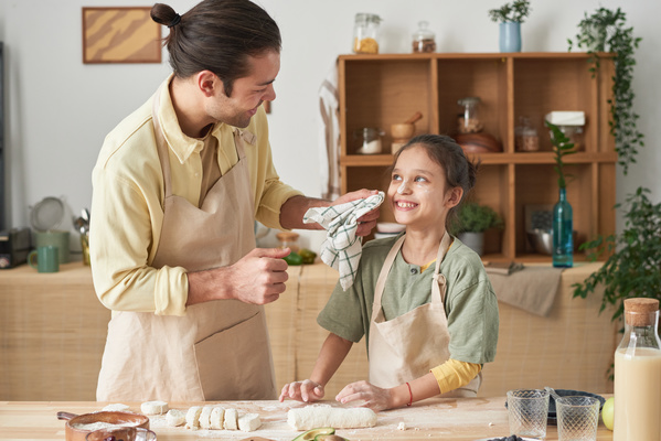A father in an apron laughing removes flour with a towel from the face of his happy daughter with her hair in a bun