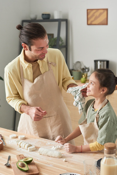 A father in an apron and with flour on his face wipes flour from the cheek of a laughing daughter with her hair in a bun