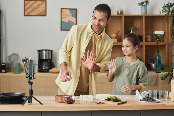 A smiling father and his daughter raising their hands in greeting while making photo on the phone which is on a tripod on the kitchen table