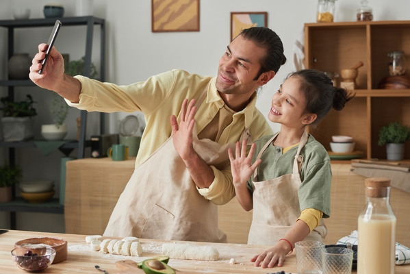 A smiling father wearing an apron takes a selfie with his daughter raising her hand in greeting