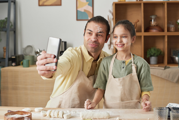 A man in an apron and his daughter with her hair in a bun taking selfies in the kitchen with flour-stained faces