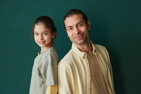 Portrait of a daughter with tidied up hair and blue eyes and a smiling father with brown eyes dressed in a yellow shirt standing against a green background