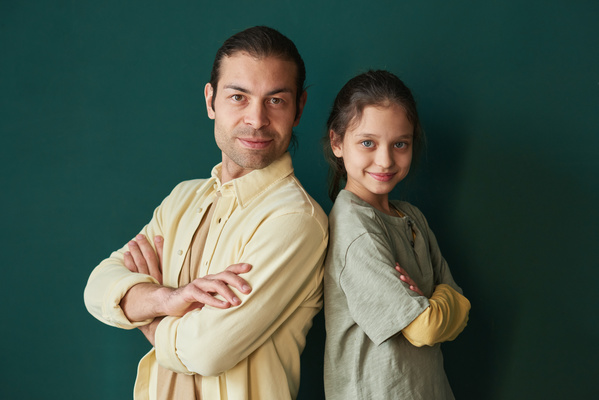 A smiling daughter with tidied up hair and blue eyes standing back to back with her father with brown eyes dressed in a yellow shirt with their arms crossed on the chests
