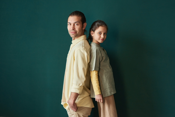 A man in a yellow shirt and his daughter pose against a dark green background standing back to back