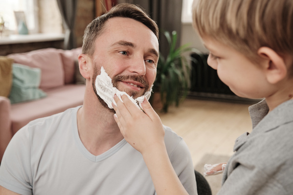 A boy with blond hair applies shaving foam to the beard of smiling father in a gray V-neck T-shirt