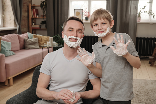 A boy with blond hair showing his hands stained with shaving foam and a father sitting in a chair have shaving foam on their faces