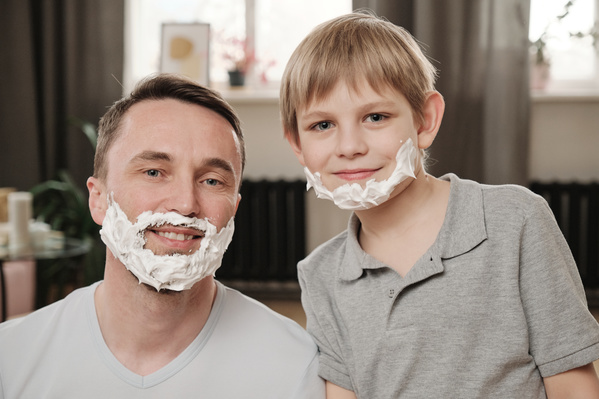 A boy with blond hair and his father dressed in gray T-shirts with shaving foam on their chins
