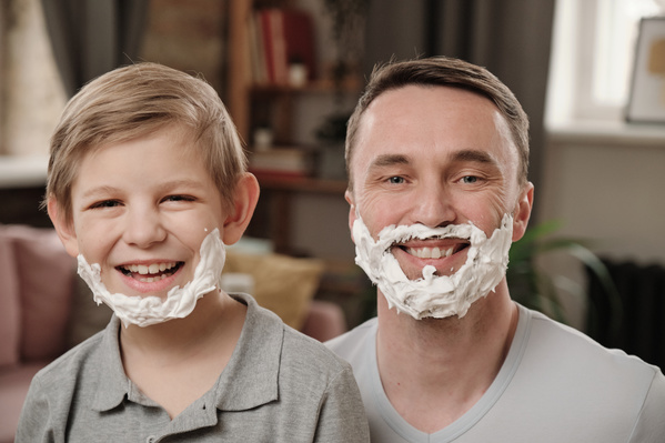 A smiling to the teeth blonde boy and his dad with shaving cream on their faces