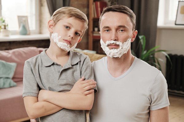 A blond son with his arms crossed on his chest and his father dressed in gray T-shirts with shaving foam on their serious faces