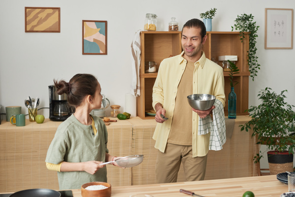 A father in a yellow shirt bringing a metal bowl and a towel to the table where his daughter standing with a sieve in her hands