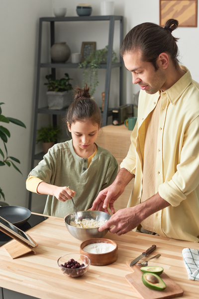 A girl with tidied up hair helps her dad dressed in a yellow shirt to mix the ingredients for the dough in a metal bowl