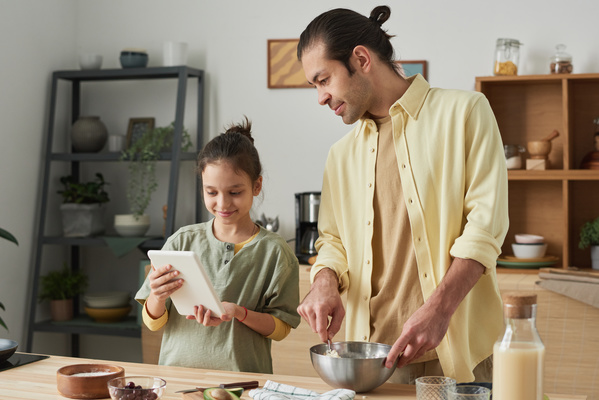 A man in a yellow shirt cooking dough in a metal bowl looks at the recipe on a white tablet held by his daughter with her hair in a bun