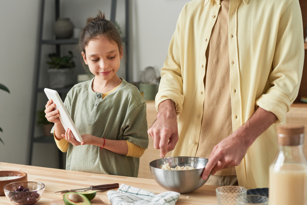 A girl with her hair in a bun and holding a white tablet in her hands watches her father mixing ingredients in a metal bowl