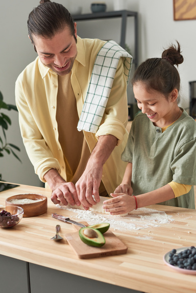 A man in a yellow shirt with a towel on his shoulder shapes a roll out of dough at the kitchen table with his daughter with tidied up hair