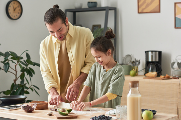 A dad in a yellow shirt and a daughter with tidied up hair smiling shaping a roll from the dough on the kitchen table