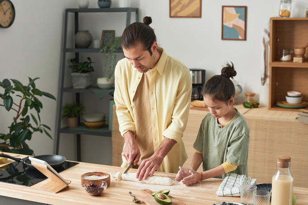 A man in a yellow shirt cutting dough into pieces and his daughter making sweet cheese pancakes out of dough pieces in the kitchen