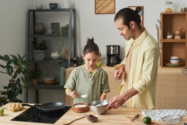 A girl with her hair in a bun adding flour to a bowl of cottage cheese held by her father in a yellow shirt