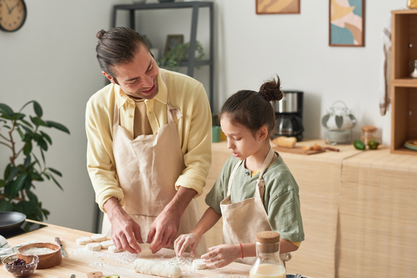 A smiling father and daughter with dark tidied up hair in a bun in light aprons shaping sweet cheese pancakes out of а dough on the kitchen table