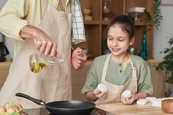 A man in light apron pouring oil on a frying pan with sweet cheese fritters that his daughter with her hair tidied up is about to fry