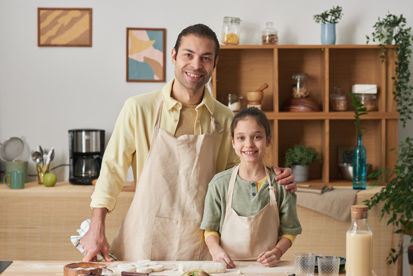 A smiling man in an apron with a towel in his hands hugging his daughters shoulder in the kitchen