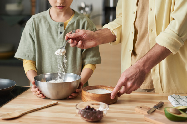 Dad adding flour with a spoon from a wooden bowl to a metal bowl that his daughter holds with both hands