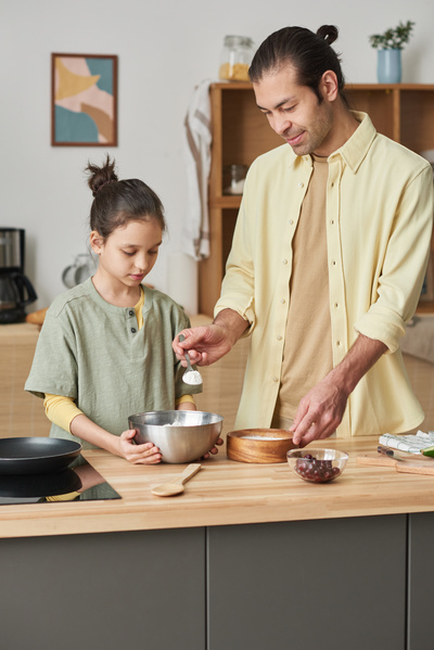 A man pouring flour with a spoon from a wooden bowl into a metal bowl that his daughter holds with both hands