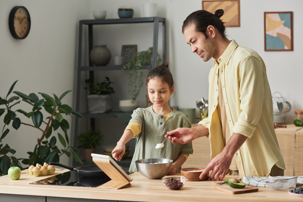 A girl with tidied up hair pointing to a recipe on a tablet to dad in a yellow shirt pouring flour into a metal bowl