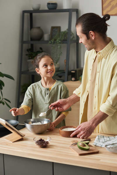 A girl with tidied up hair looks at her dad in a yellow shirt adding flour to a metal bowl and pointing to the recipe in the tablet
