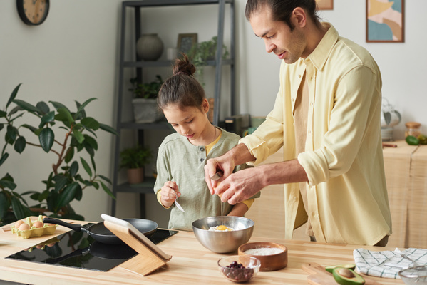 A girl with her hair tidied up watches her father in a yellow shirt breaking an egg into a metal bowl with flour