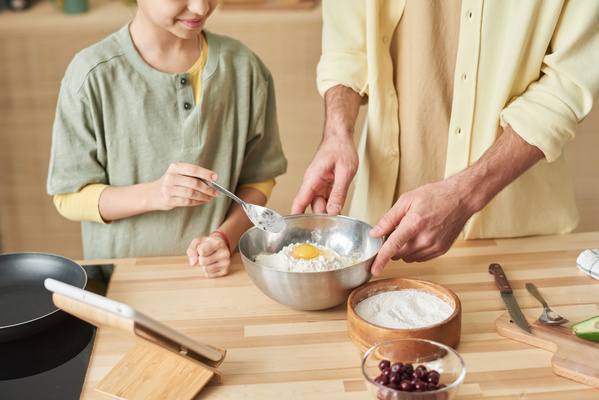 A girl is about to stir the dough with a spoon in a metal bowl held by her father in a yellow shirt
