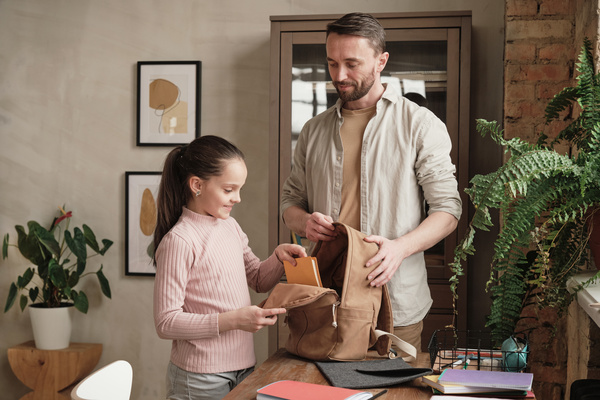 A man in a light shirt over a T-shirt looks at his daughter with tidied up hair putting a book in a school bag